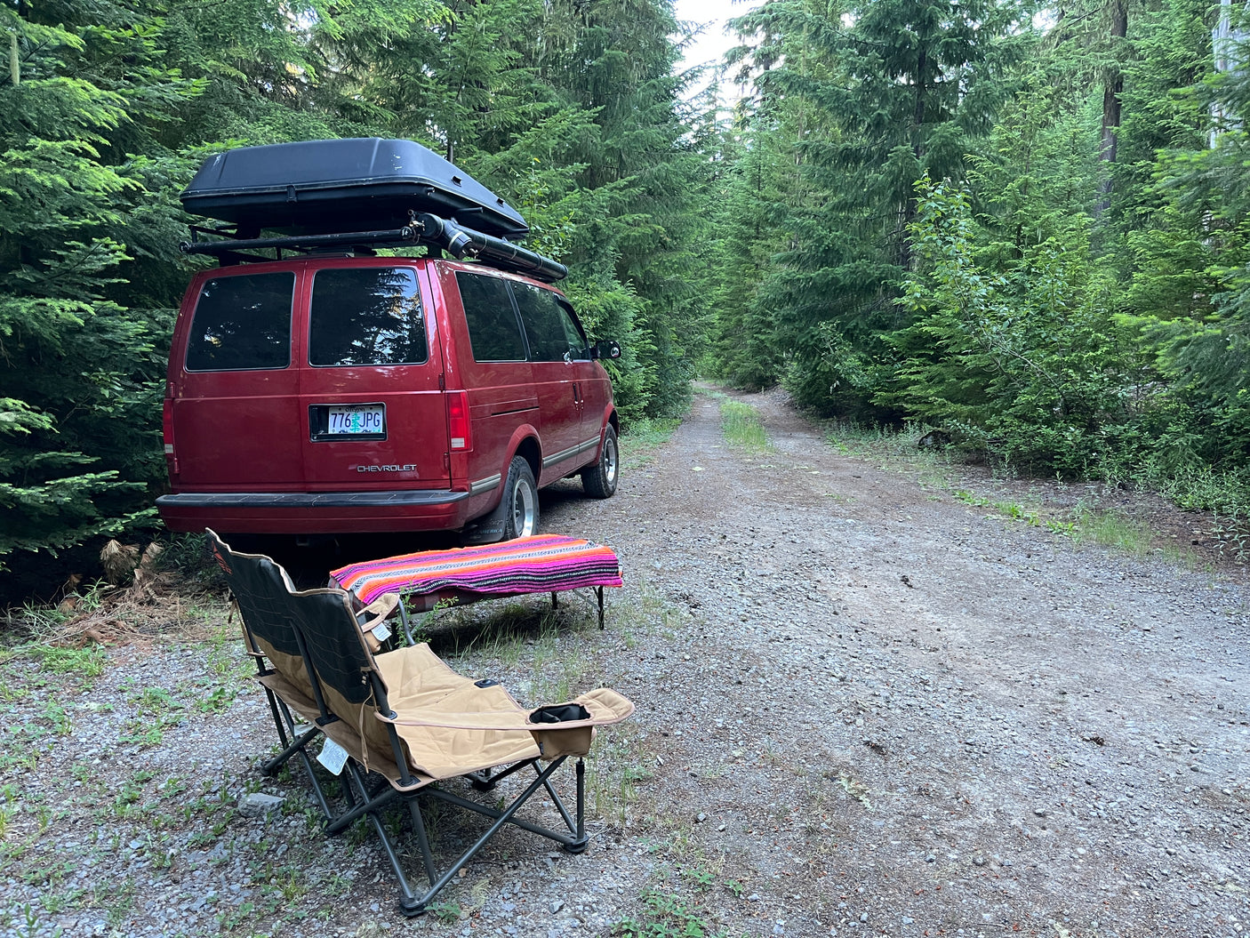 Red camper van with roof box on a forest road, chairs & table set up for off-grid RV rental camping.