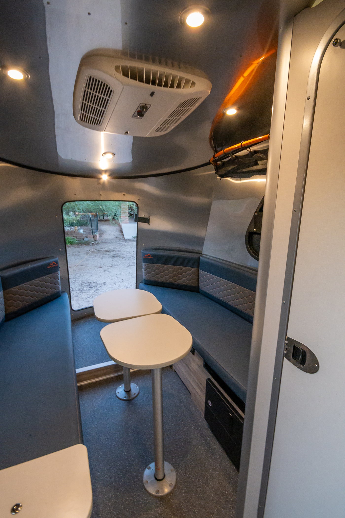 Cozy RV rental interior: a compact dining area with blue bench seating, dual tables, and AC. View outdoors from the rear door. — photo 19