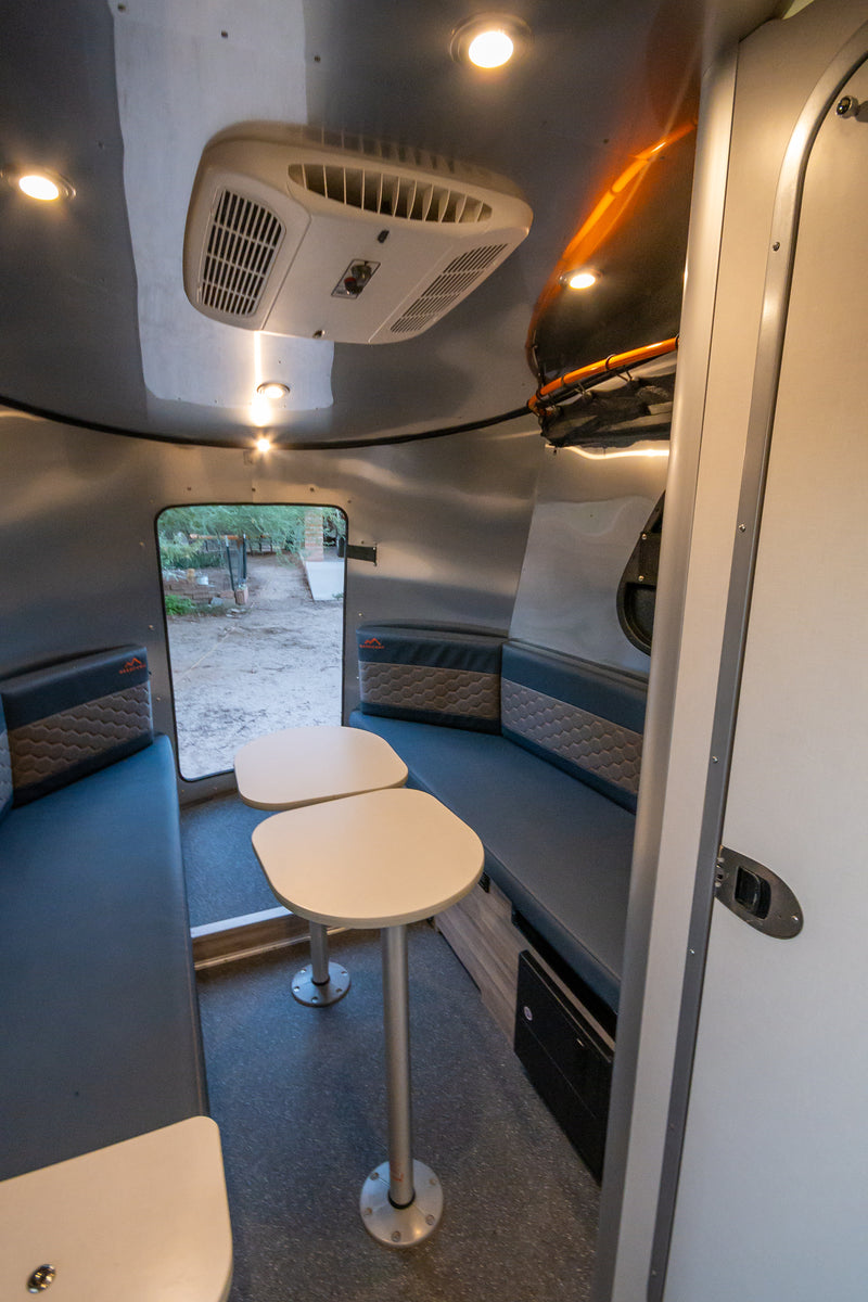 Cozy RV rental interior: a compact dining area with blue bench seating, dual tables, and AC. View outdoors from the rear door. — photo 19
