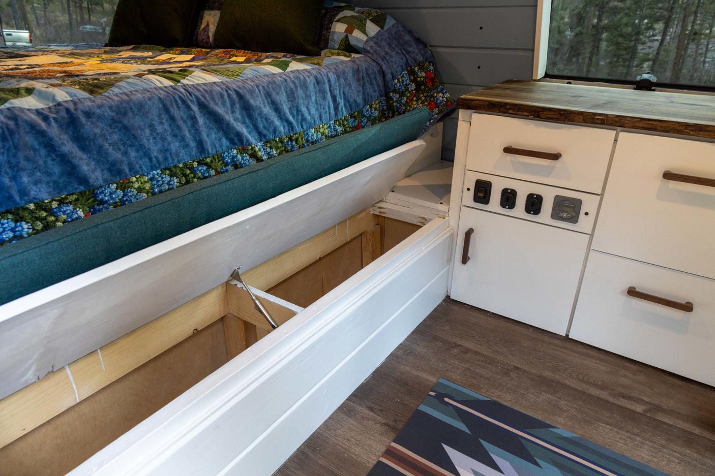 Clever storage in this cozy RV rental! An under-bed compartment is open, beside white cabinets with controls.