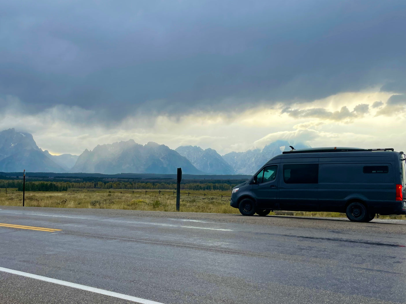 An rv rental van with a surfboard, parked on a wet road. Majestic mountains and cloudy sky are in the background.