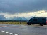 An rv rental van with a surfboard, parked on a wet road. Majestic mountains and cloudy sky are in the background.