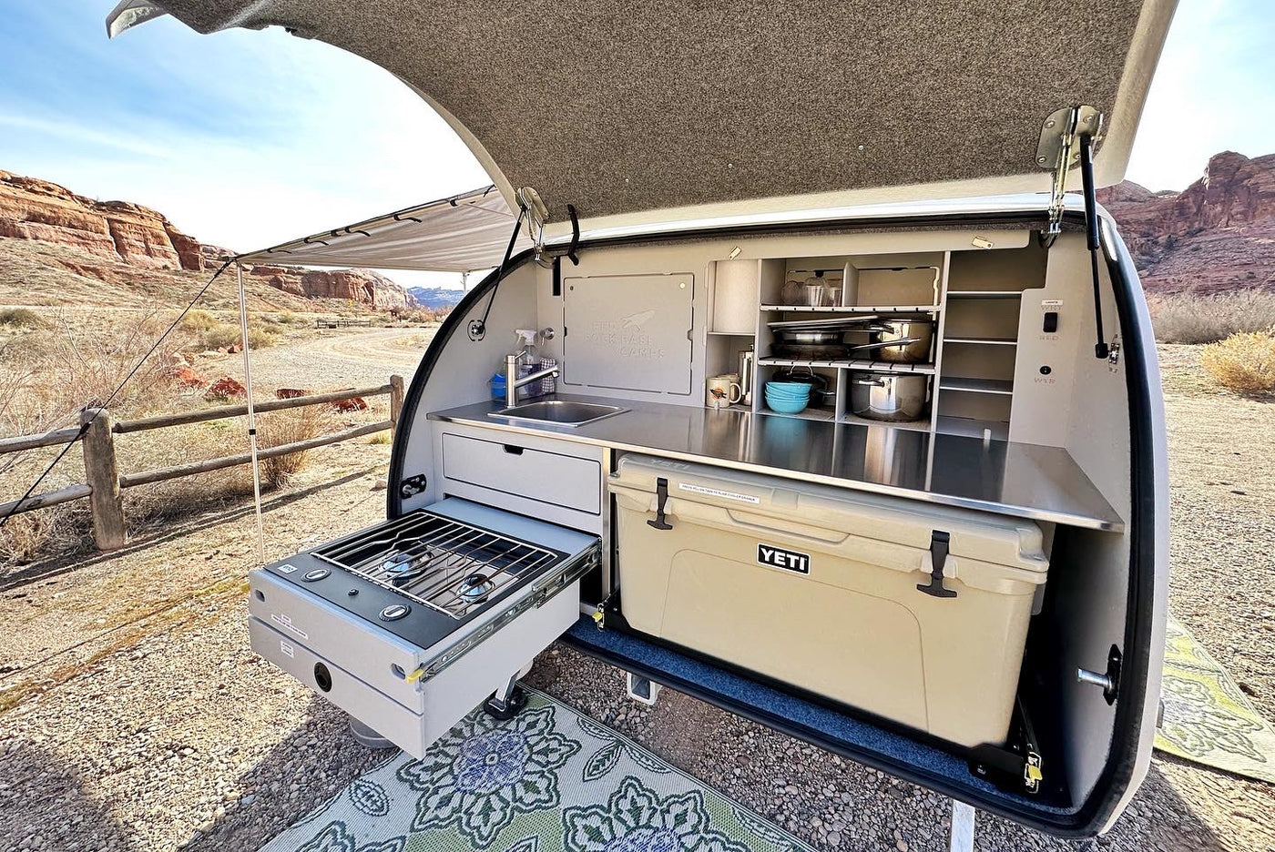 Outdoor kitchen of an RV rental teardrop trailer in a desert setting. Features stove, sink, cooler, & organized storage for camping. — photo 13