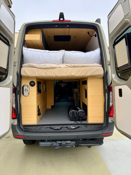 Inside an RV rental: rear view of a comfortable camper van bed, custom bamboo storage, and open cargo area.