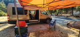 Tan Sprinter rv rental with an orange awning & open side for camp. Shows a blue chair, red cooler, and mat for outdoor living.