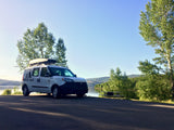 White 'notre vans' RV rental with rooftop cargo box, parked by a tranquil lake, surrounded by trees and mountains.