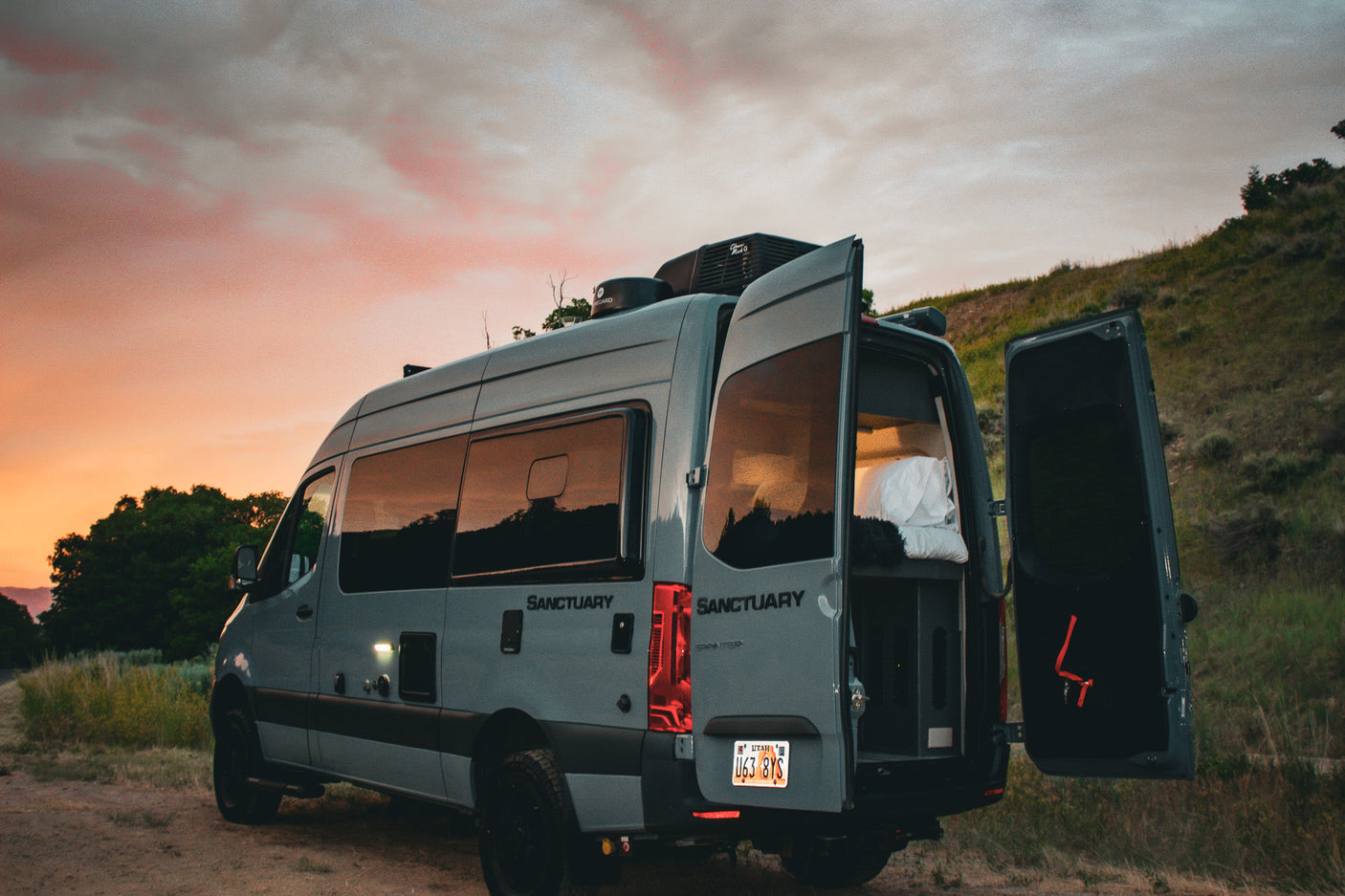 Gray camper van RV rental parked on a dirt road at sunset, rear doors open revealing the cozy interior. Ready for adventure!