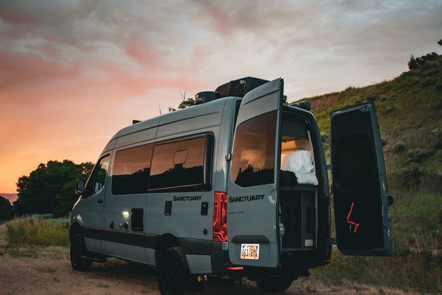 Gray camper van RV rental parked on a dirt road at sunset, rear doors open revealing the cozy interior. Ready for adventure!