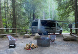 A forest RV rental campsite with a camper van, fire pit, chairs, and bikes, ready for outdoor living and remote work.