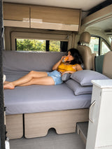A woman relaxes on a bed inside a modern RV rental camper van, enjoying a drink. Perfect for comfortable travel.