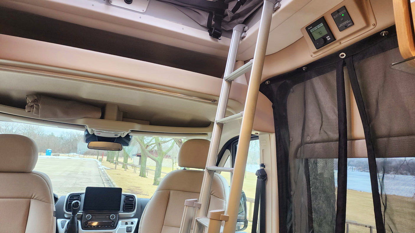 Inside an RV rental: View from the rear, showing front seats, overhead storage, ladder to bunk, and control panel. Lake seen through screen.