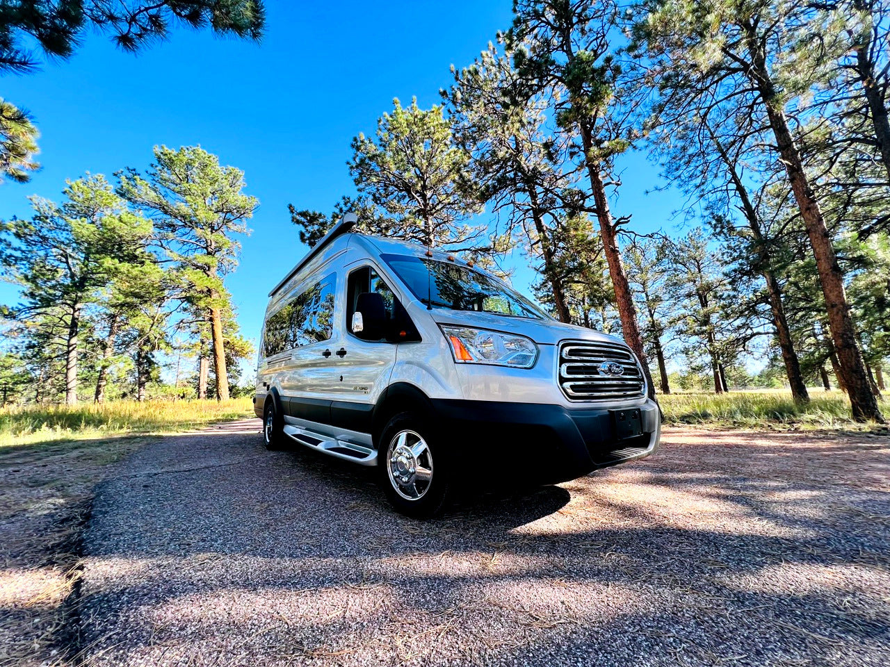 Silver camper van RV rental, parked on a gravel road among tall pines under a bright blue sky. Ready for your next outdoor adventure.