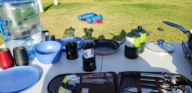 Camping essentials for an RV rental adventure: water, cooking gear, lanterns, and utensils laid out on a table outdoors.