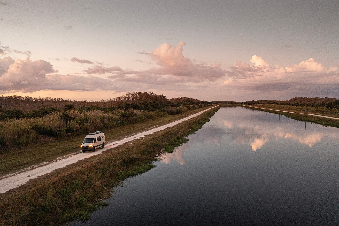 An RV rental camper van explores a scenic dirt road alongside a peaceful canal at sunset, reflecting clouds. — photo 8