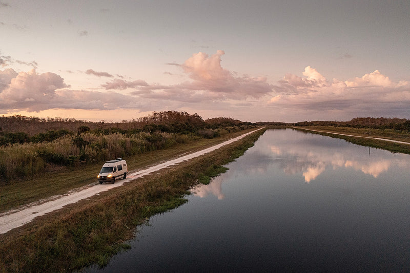 An RV rental camper van explores a scenic dirt road alongside a peaceful canal at sunset, reflecting clouds. — photo 8