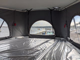 Inside an RV rental pop-top, a bed with plastic cover overlooks a tranquil harbor with boats.