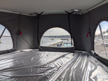 Inside an RV rental pop-top, a bed with plastic cover overlooks a tranquil harbor with boats.