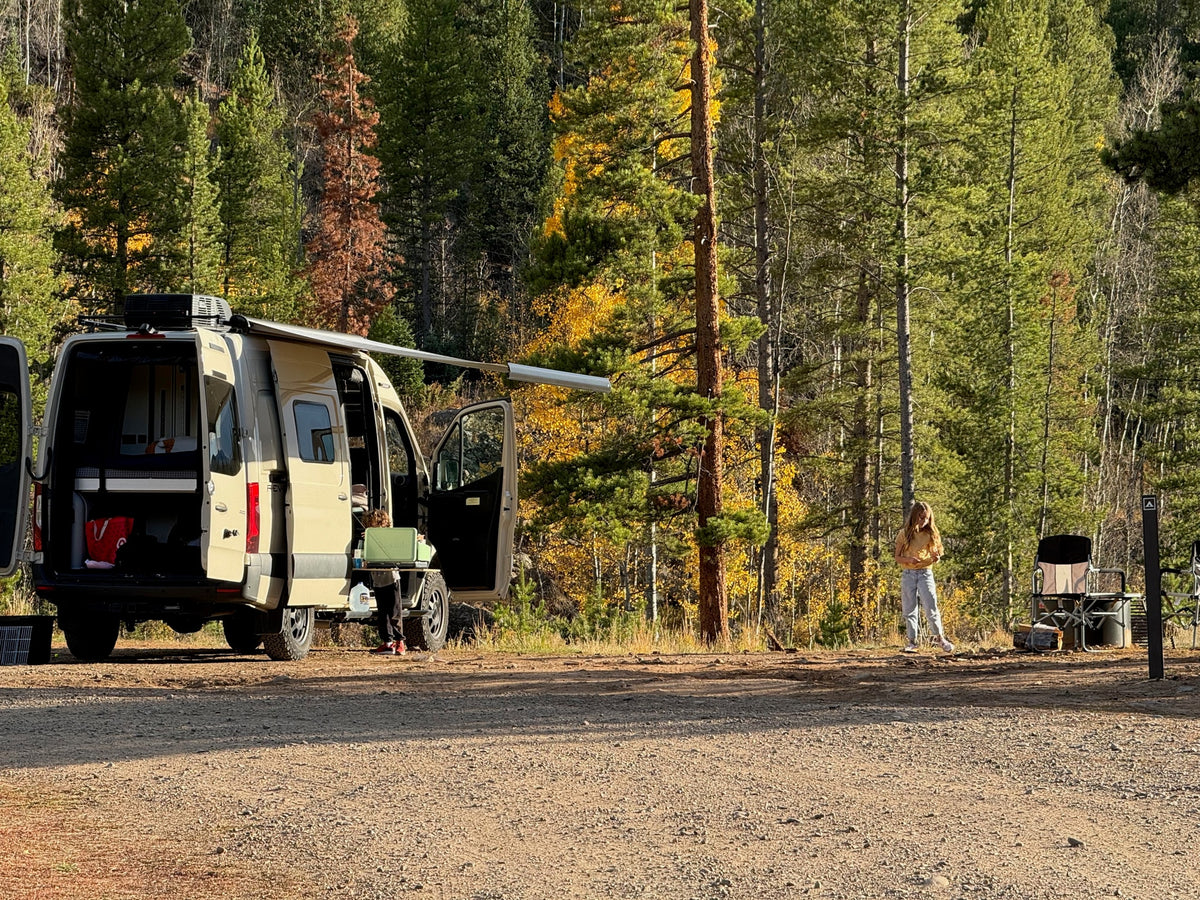 A person stands by their RV rental camper van in a vibrant fall forest campsite.