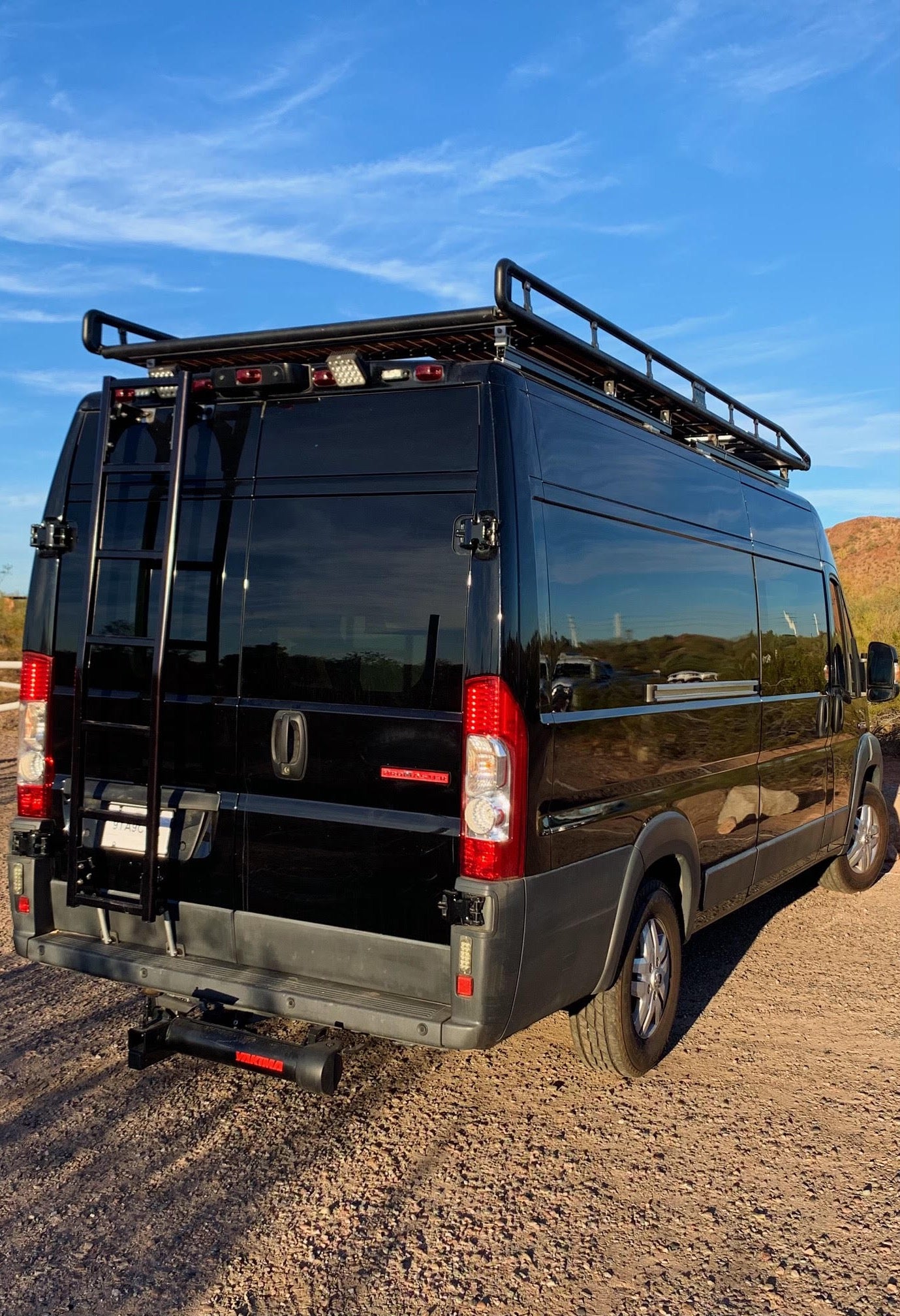 Black camper van RV rental, featuring a roof rack and rear ladder, parked on a dirt road in a desert setting.