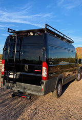 Black camper van RV rental, featuring a roof rack and rear ladder, parked on a dirt road in a desert setting.
