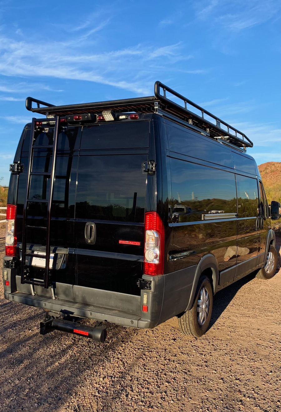 Black camper van RV rental, featuring a roof rack and rear ladder, parked on a dirt road in a desert setting.