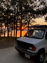 White Ford RV rental van parked by a lake at sunset. Golden light reflects on the water through silhouetted trees. Ideal road trip!