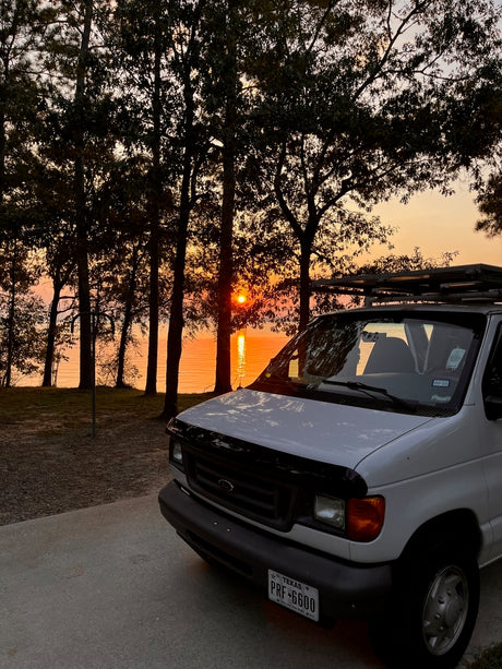 White Ford RV rental van parked by a lake at sunset. Golden light reflects on the water through silhouetted trees. Ideal road trip!