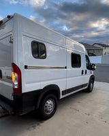 White Ram ProMaster camper van for RV rental, with custom side windows, parked outdoors under a cloudy sky.