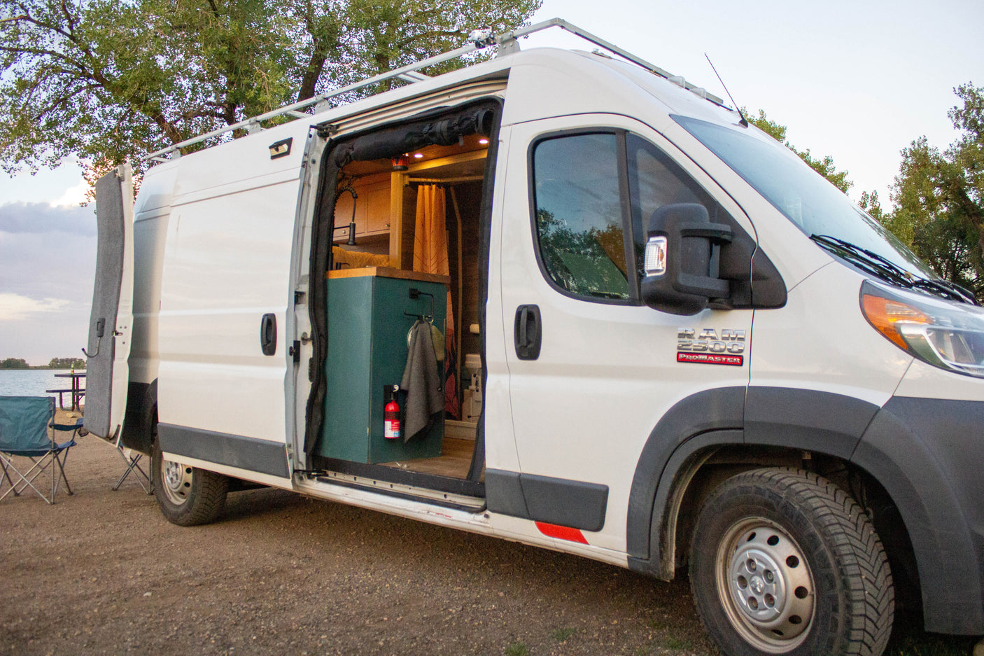 White Ram ProMaster camper van with open side door at a scenic lakeside campsite, ready for your next RV rental.