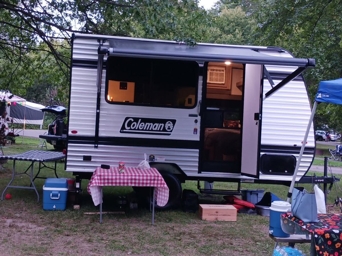  with houses behind it.|A white Coleman travel trailer (RV rental) is parked in a pine forest campsite with a black and white Great Dane dog nearby.|A white GMC pickup is hitched to a Coleman RV rental travel trailer with interior lights on — photo 5