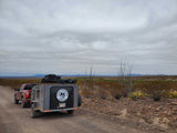 Off-road RV rental from Texas Adventure Rentals, hitched to a truck on a rugged desert road under a cloudy sky.
