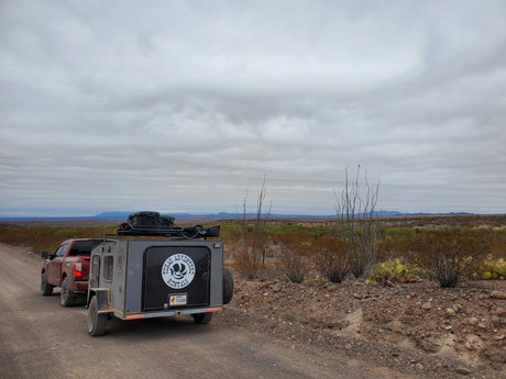Off-road RV rental from Texas Adventure Rentals, hitched to a truck on a rugged desert road under a cloudy sky.