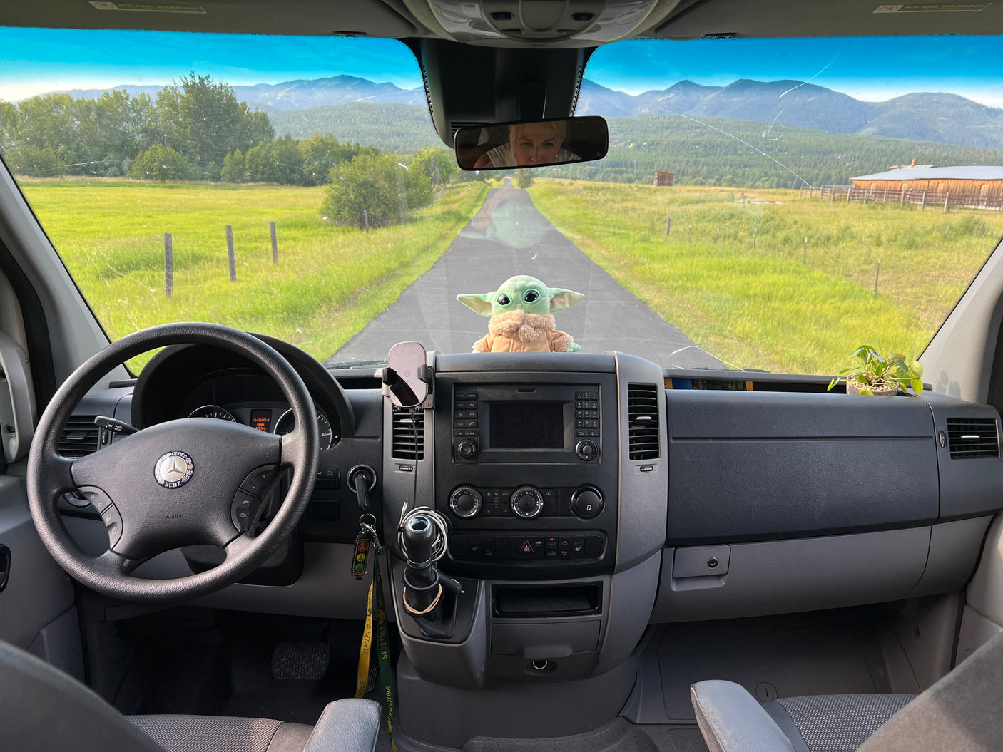 Inside an rv rental van, a Grogu plush watches a scenic road to mountains. Driver's reflection and cracked windshield are visible.