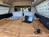 Inside an RV rental: a cozy truck camper bed with pillows, patterned curtains, and a laptop charging for travel.