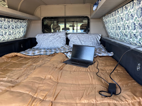 Inside an RV rental: a cozy truck camper bed with pillows, patterned curtains, and a laptop charging for travel.