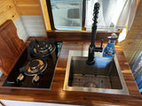 Modern RV rental kitchenette: sleek wood counters, subway tile backsplash, two-burner stove, and stainless steel sink.