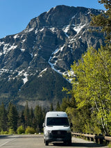 White RV rental van parked on a mountain road, majestic snow-capped peak and lush green forest filling the background.