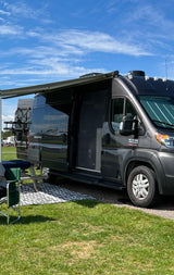 Dark gray RV rental with extended awning and open screen door, parked on grass. Ideal for a summer camping trip.