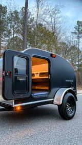 Cozy black teardrop camper, door open revealing wood interior & blue mattress. Perfect for rv rental adventures.
