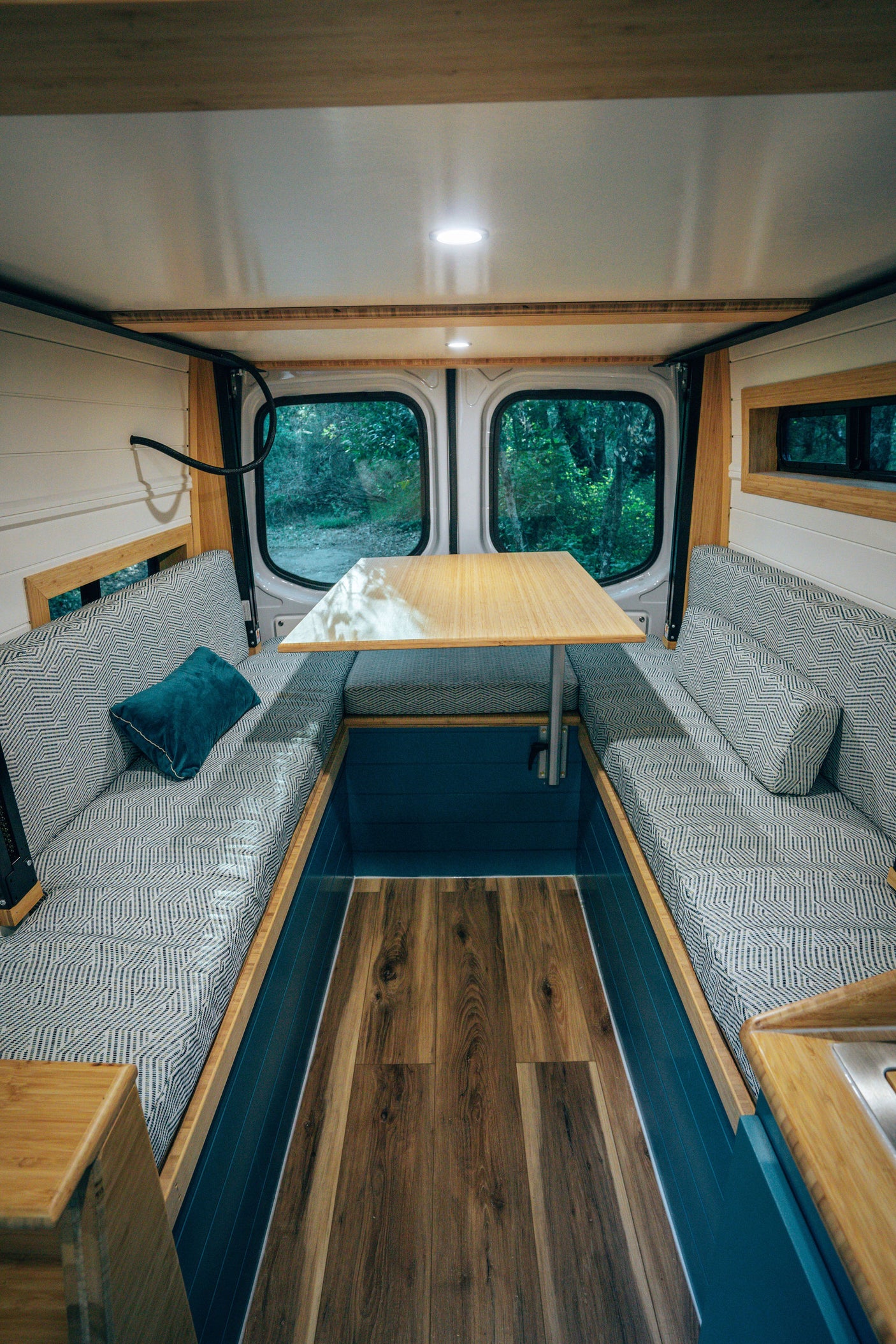 Cozy rv rental interior: wood-paneled van dining area with patterned cushions, overhead light, and forest views.