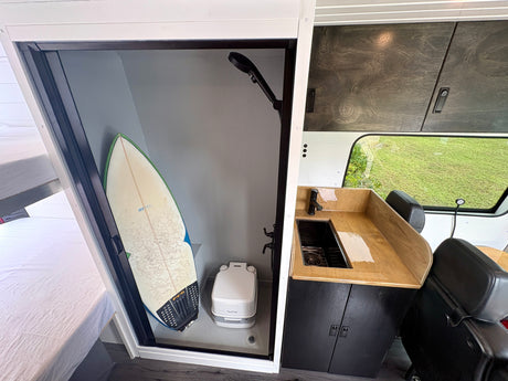 This RV rental showcases a compact interior with a shower, toilet, surfboard, kitchenette, and sleeping area.