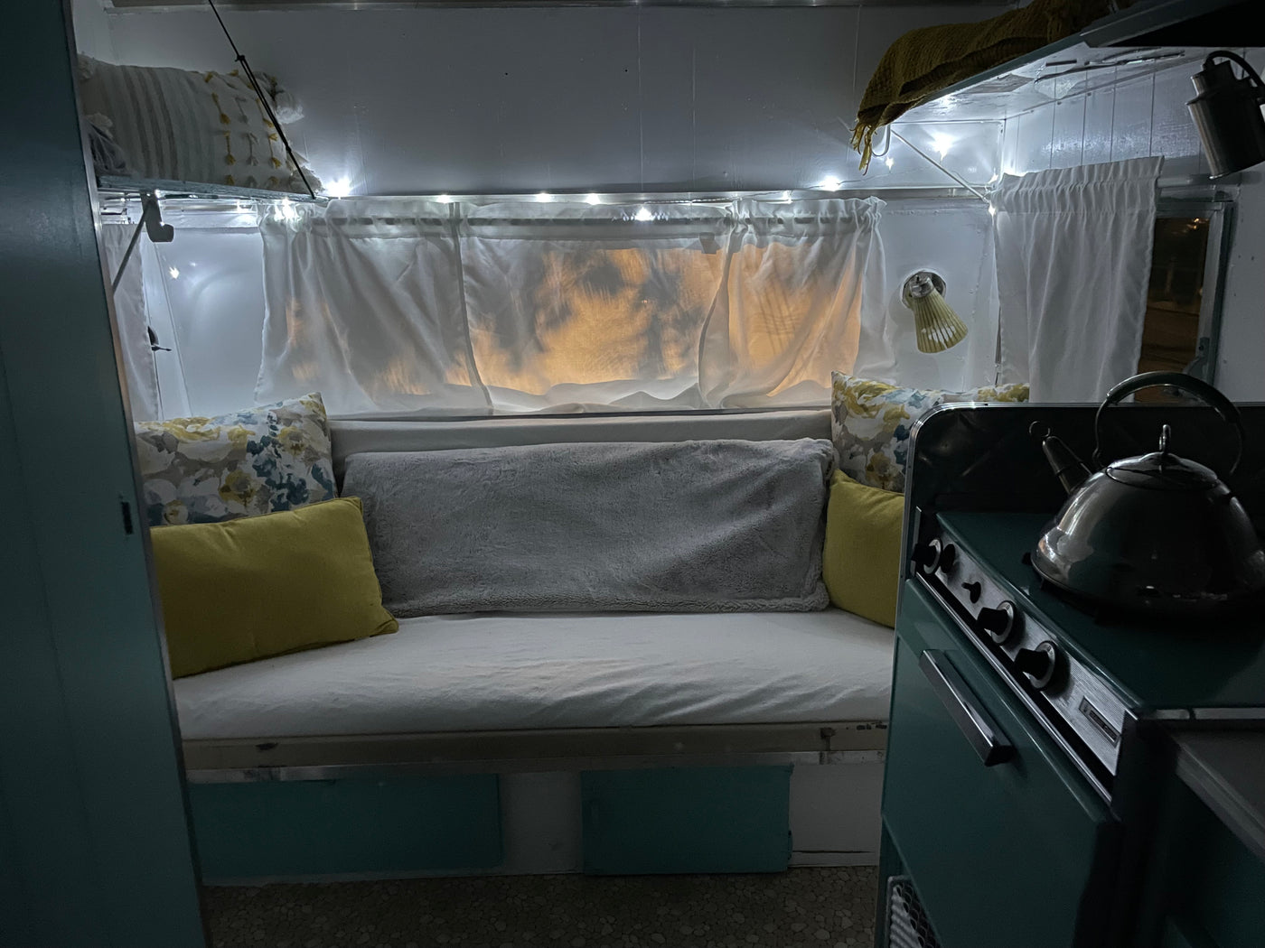 Inviting RV rental interior: a comfy bed with pillows, glowing string lights, and a compact kitchen stove with a kettle. — photo 11