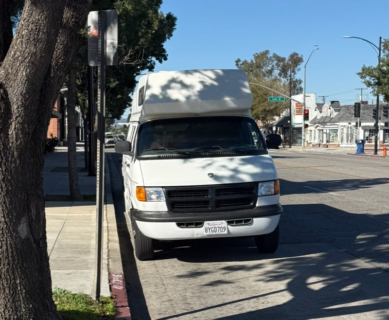  parked on a city street at night. Its high roof offers extra space for travel.|White Class B rv rental parked in a sunny lot next to a grey Honda Civic. Trees visible in the background.|White camper van — photo 3