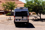 An RV rental, a silver Thor Tellaro camper van, is parked in a sunny desert campground with red rock formations behind.