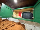 RV rental interior: overhead kitchen cabinet with dishes, tray, bag, and a cozy bed below, offering compact storage.