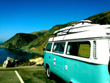 Teal VW camper van rental with boards, on a scenic coastal road overlooking a bridge.