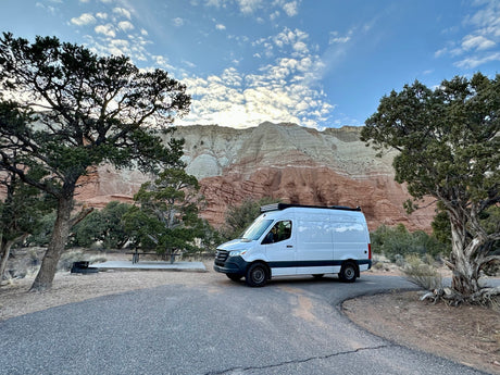 White RV rental van at a scenic campsite, framed by layered mountains under a cloudy sky.