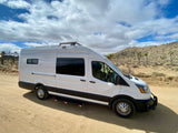 White RV rental van, customized for adventure, parked on a desert road with Joshua trees under a cloudy sky.