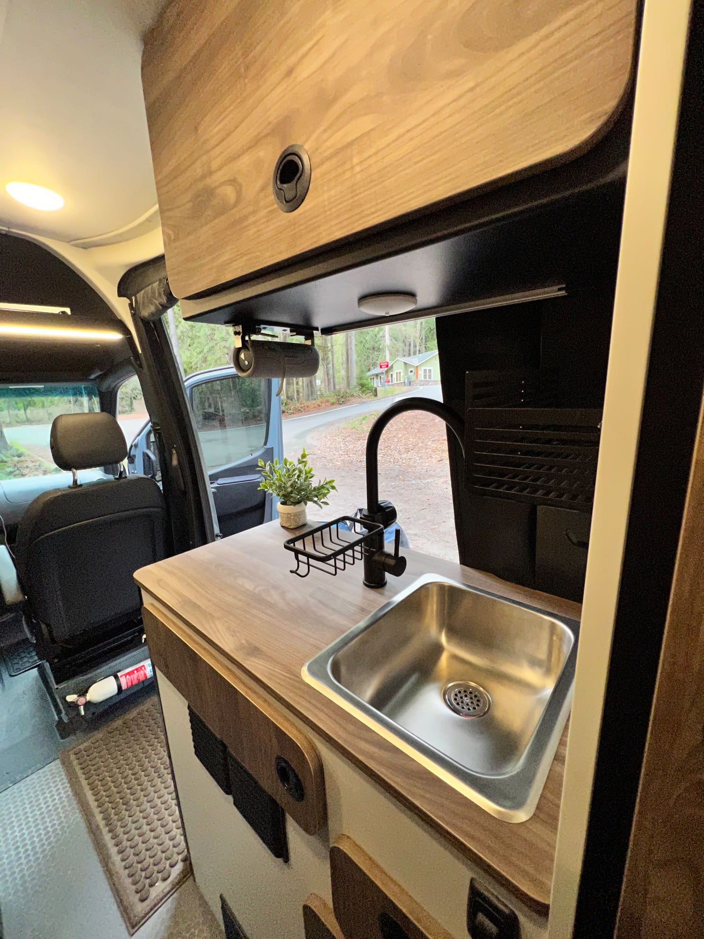 Modern RV rental interior: a sleek kitchen area with stainless steel sink, wood-look counter, and storage. Open door view to nature.