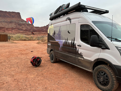 Adventure-ready RV rental van with paraglider, kayak, and unicycle in a scenic desert landscape.
